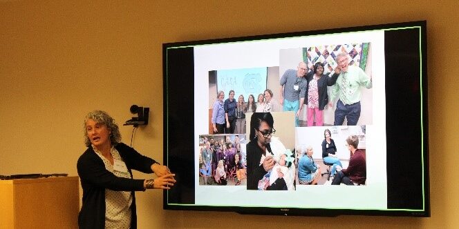 Melinda Ramage, a white woman with grey hair in a black cardigan, gestures toward a screen of photos in a conference room